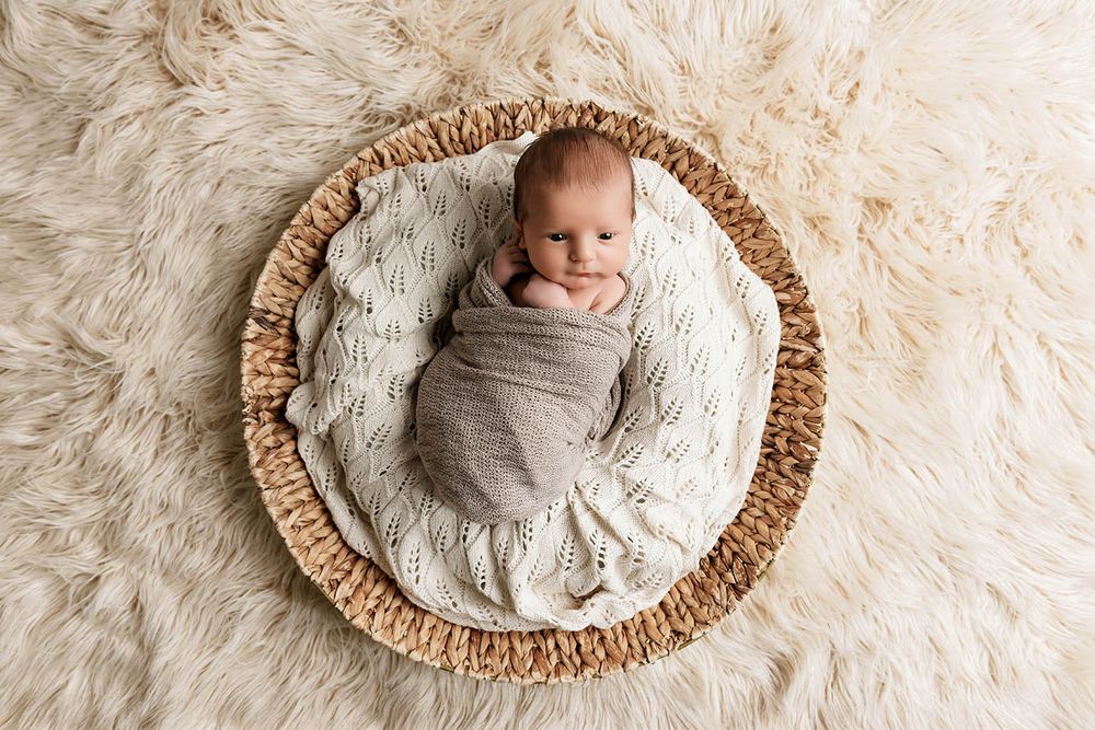 Swaddled newborn baby resting peacefully in a woven basket on a soft cream fluffy rug.