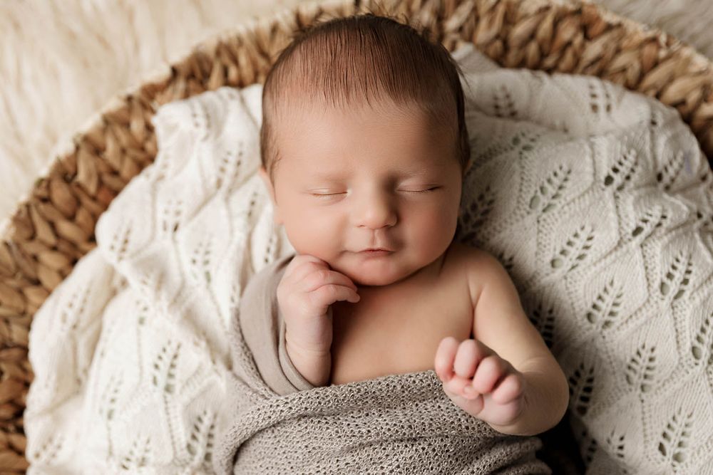 Peaceful newborn baby sleeping in a wicker basket wrapped in a soft gray knit blanket.