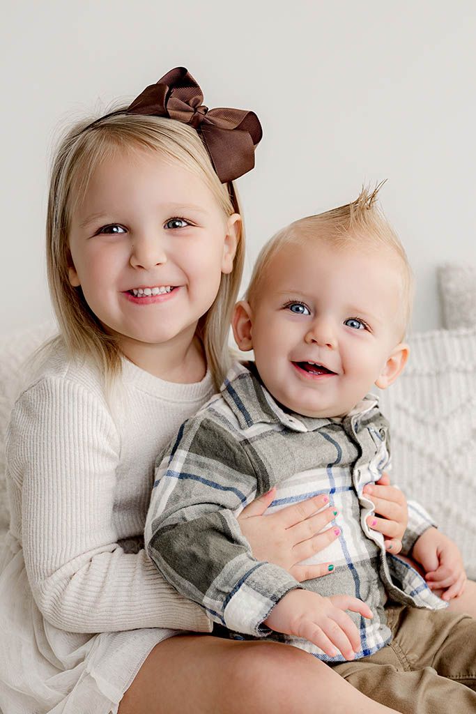Smiling toddler girl with brown bow hugging happy baby boy in plaid shirt portrait.