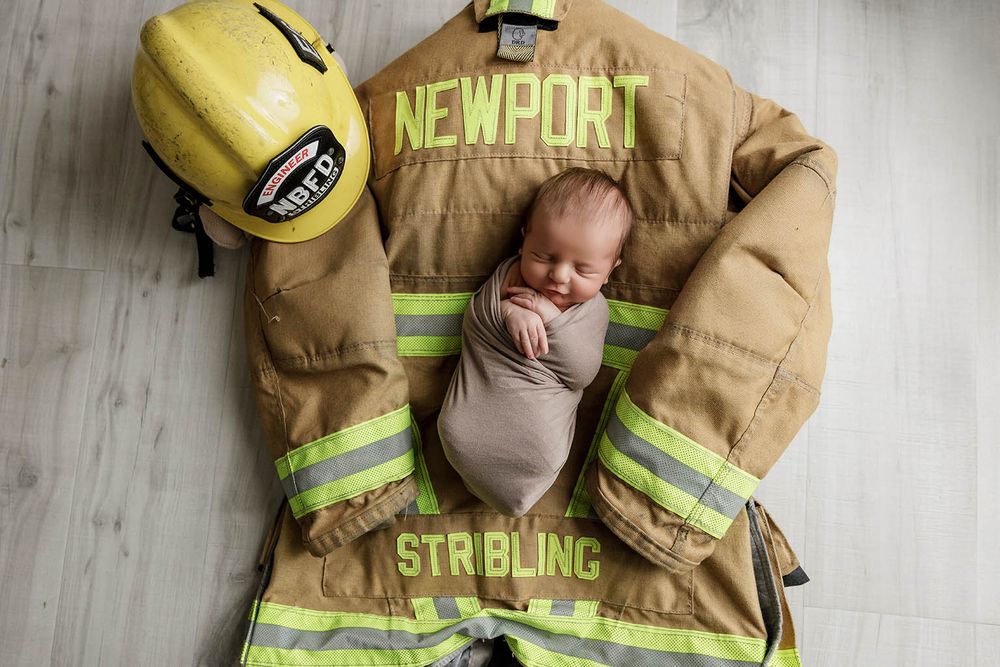 Sleeping newborn baby swaddled in gray wrap resting on Newport Stribling firefighter turnout gear.