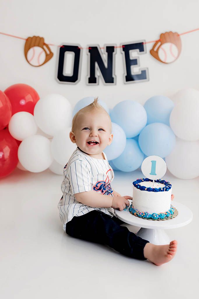 Baby boy in baseball jersey celebrates first birthday with cake, balloons, and ONE banner at a cake smash photo session.
