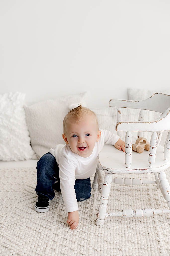 Smiling baby in white shirt and jeans crawling near a vintage white chair on a textured white rug.