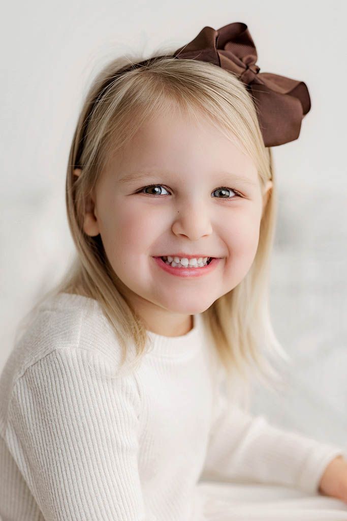Smiling blonde toddler girl wearing a white outfit and brown satin bow headband against a white background.