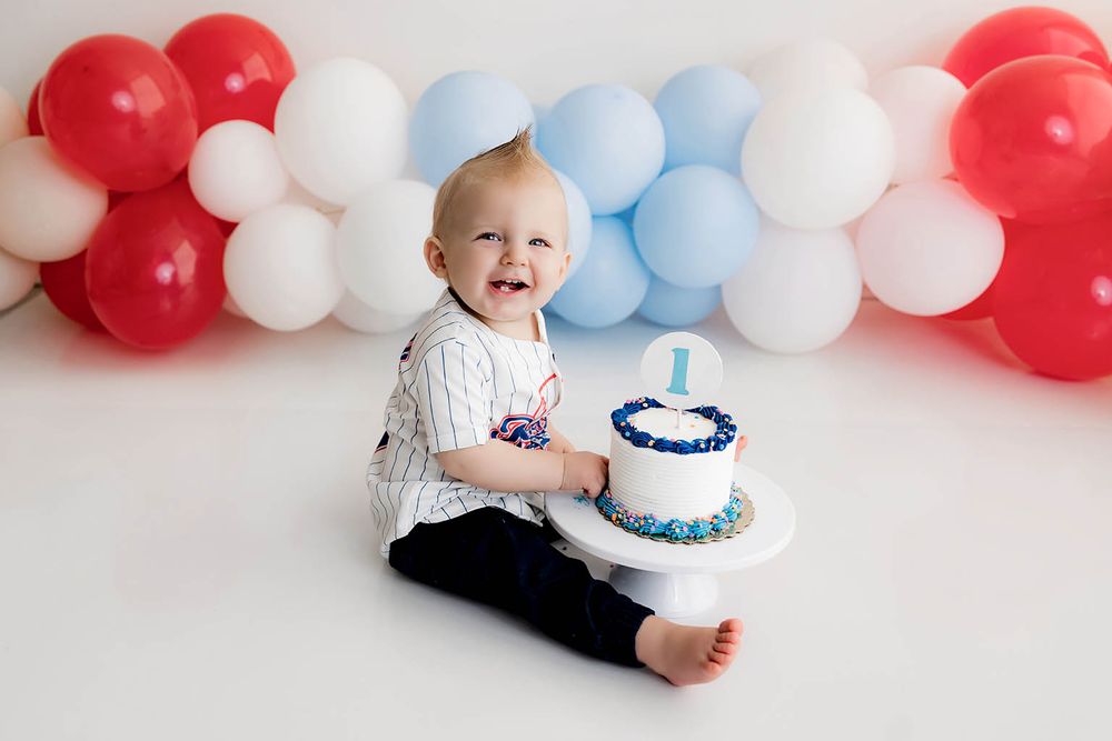 Happy baby celebrates first birthday smashing a blue and white cake surrounded by red and blue balloons.