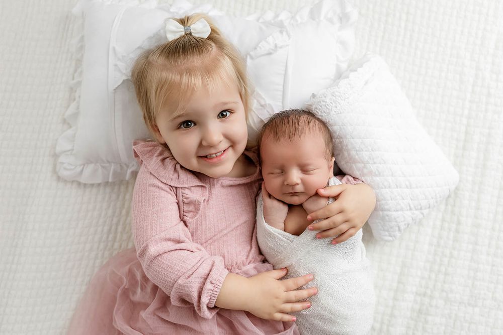 Young blonde toddler girl in pink dress smiles while gently holding sleeping newborn baby wrapped in white on a soft bed.