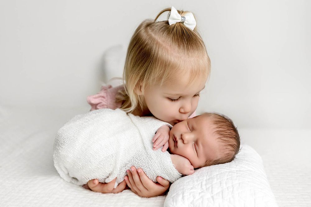Young toddler girl with bow gently kissing sleeping newborn baby wrapped in white swaddle.