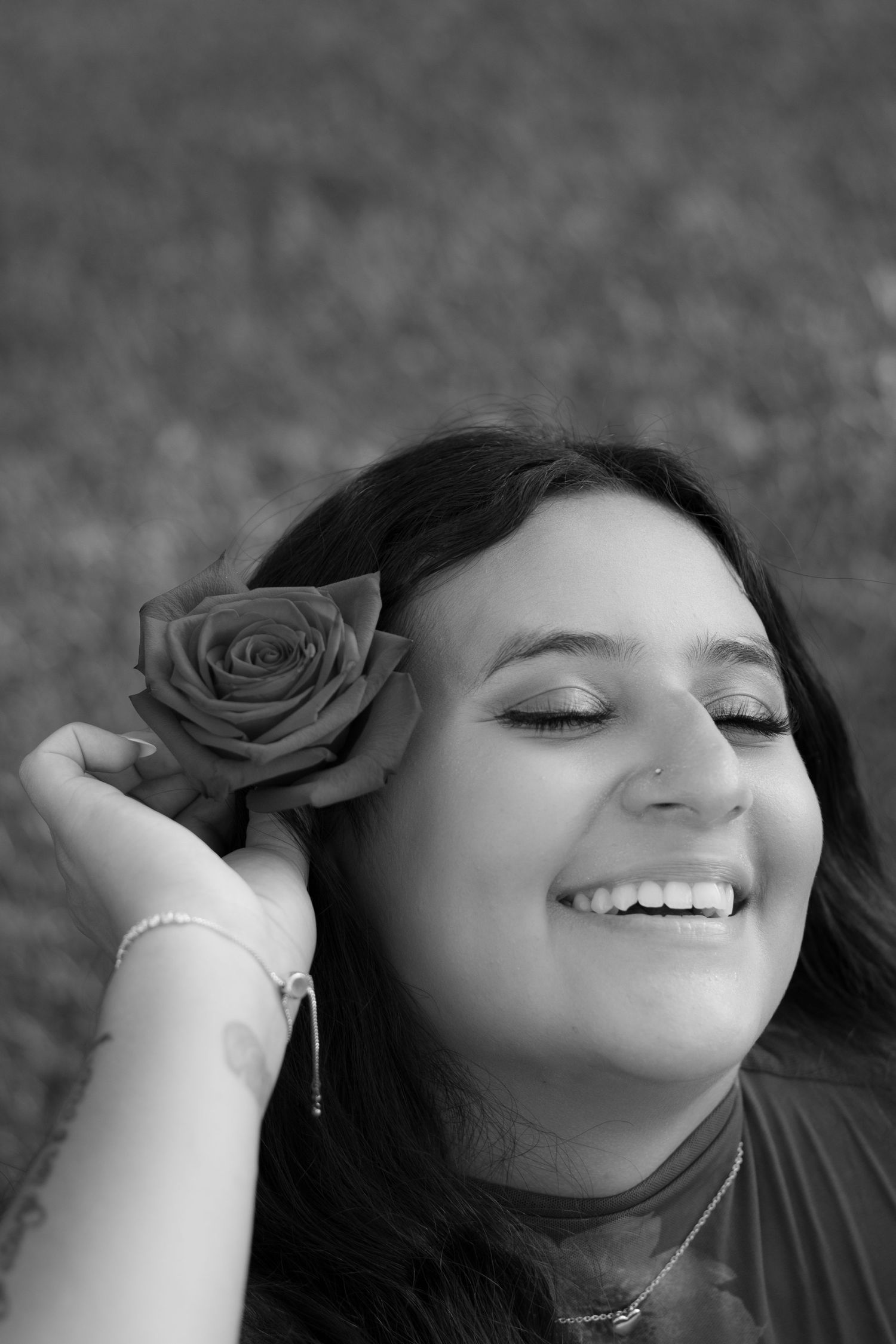Black and white portrait of a smiling woman with eyes closed holding a rose behind her ear outdoors.