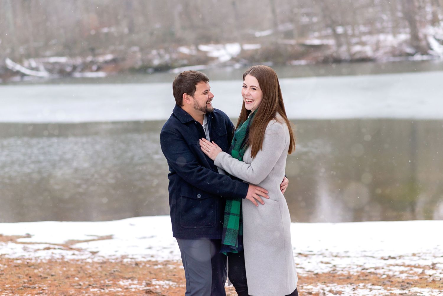 Couple embracing and smiling by a snowy lakeside in winter, wearing coats in a romantic outdoor setting.
