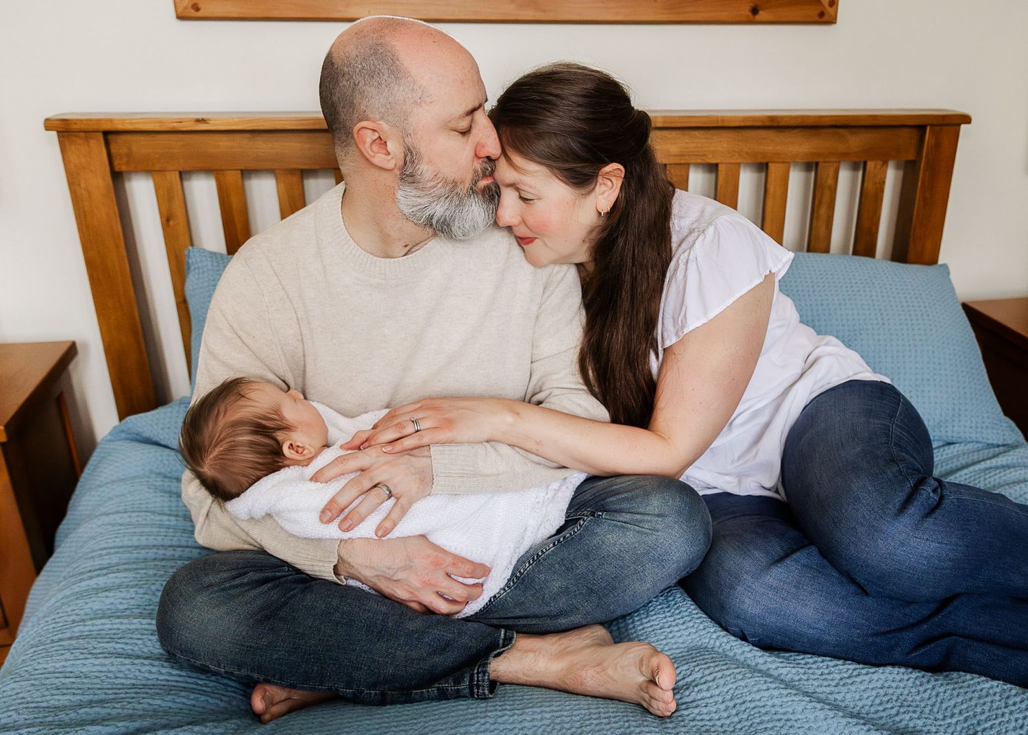 A Bristol couple sit on their bed. Dad kisses mum's forehead as he holds their newborn baby during a relaxed photoshoot at home.