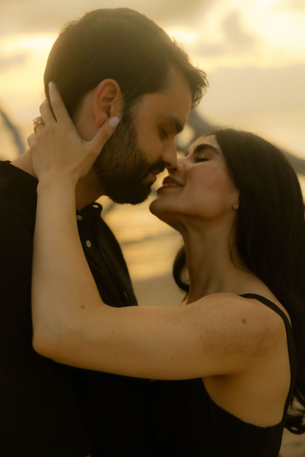 Romantic couple sharing an intimate almost-kiss at golden sunset, woman with dark hair in black dress embracing bearded man.