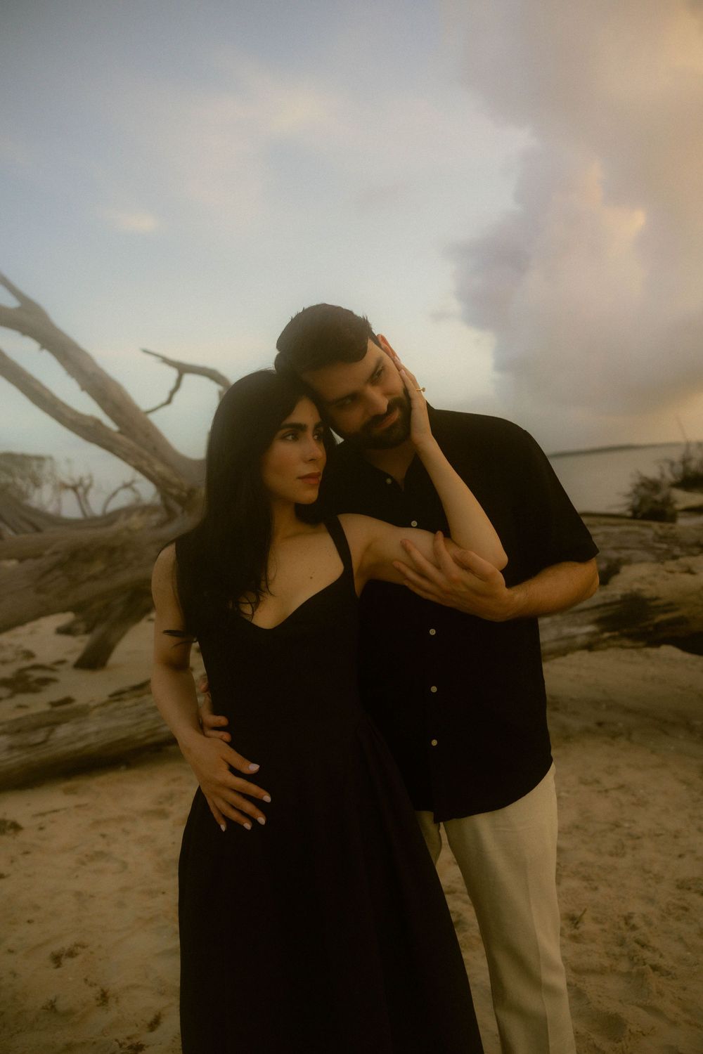 Couple in matching black outfits embracing on a sandy beach at dusk with driftwood in the background.