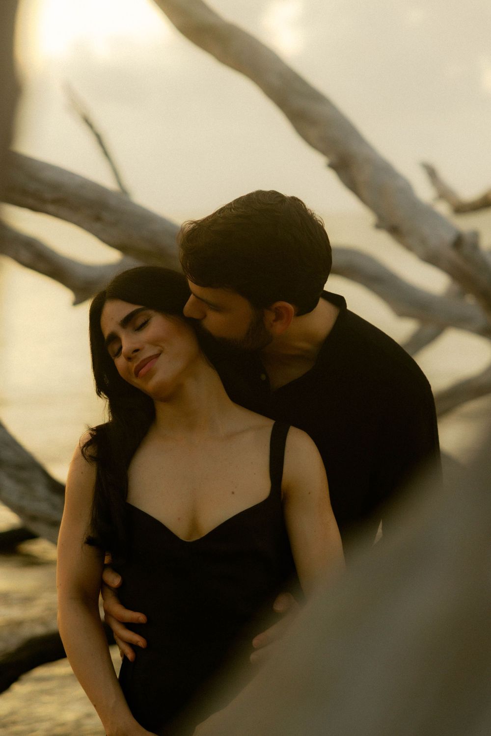 Couple in black embracing among driftwood at golden sunset on beach.