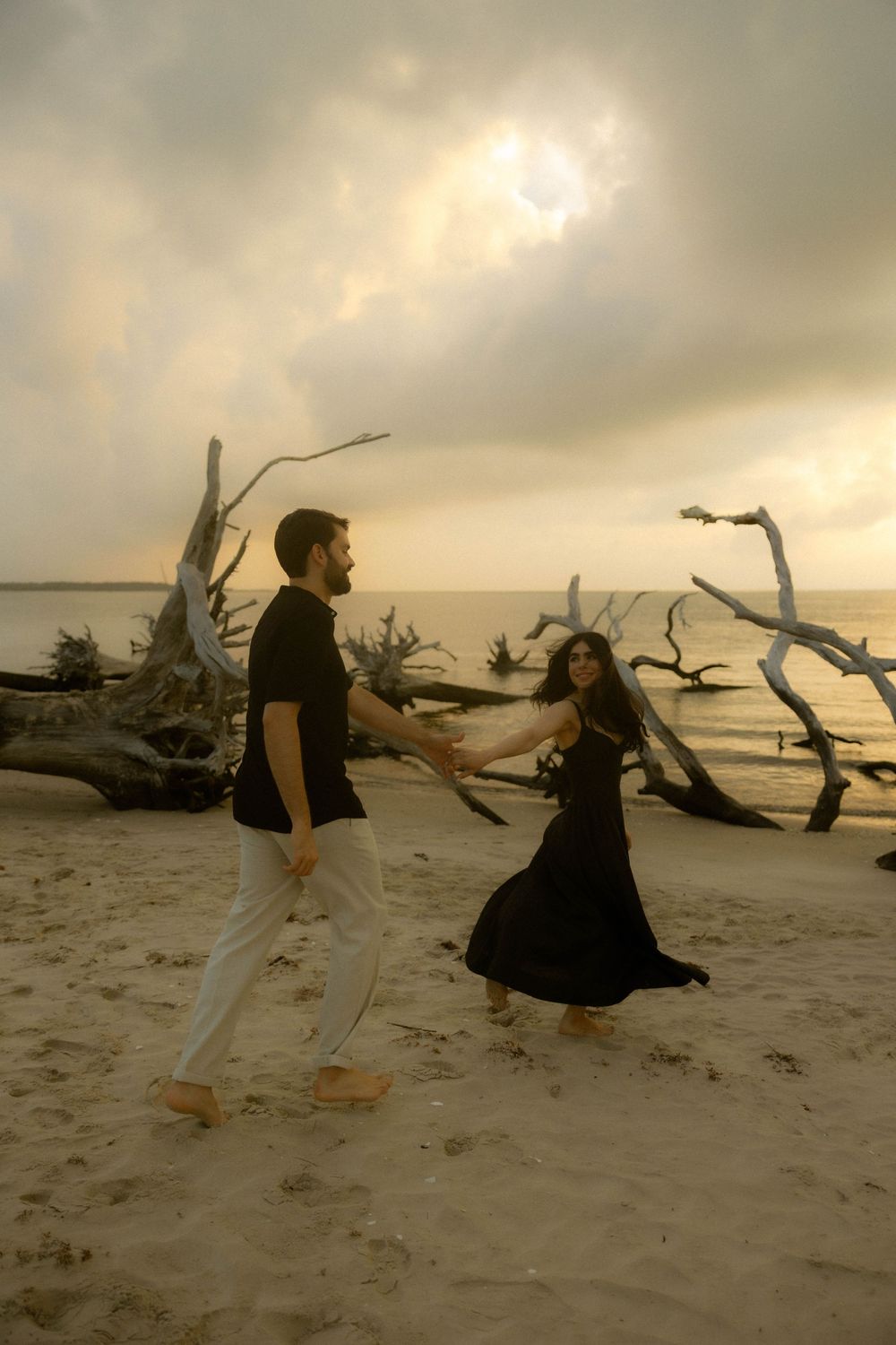 Couple dancing joyfully on sandy beach at sunset with dramatic driftwood and cloudy sky backdrop.