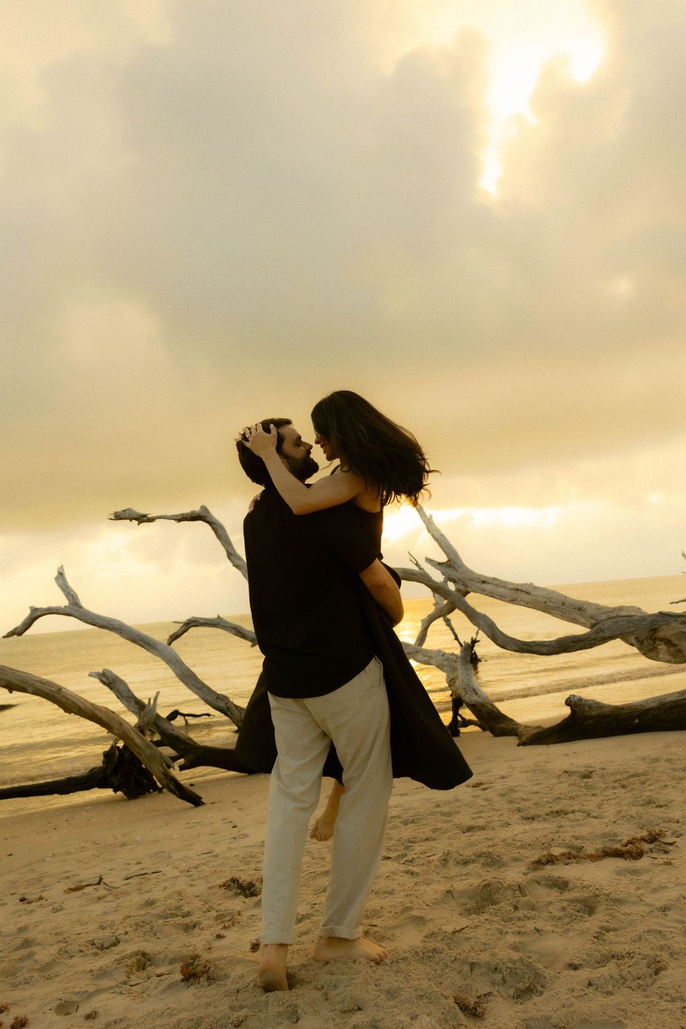 Man lifting woman in romantic embrace on beach at golden hour with weathered driftwood and ocean behind them.
