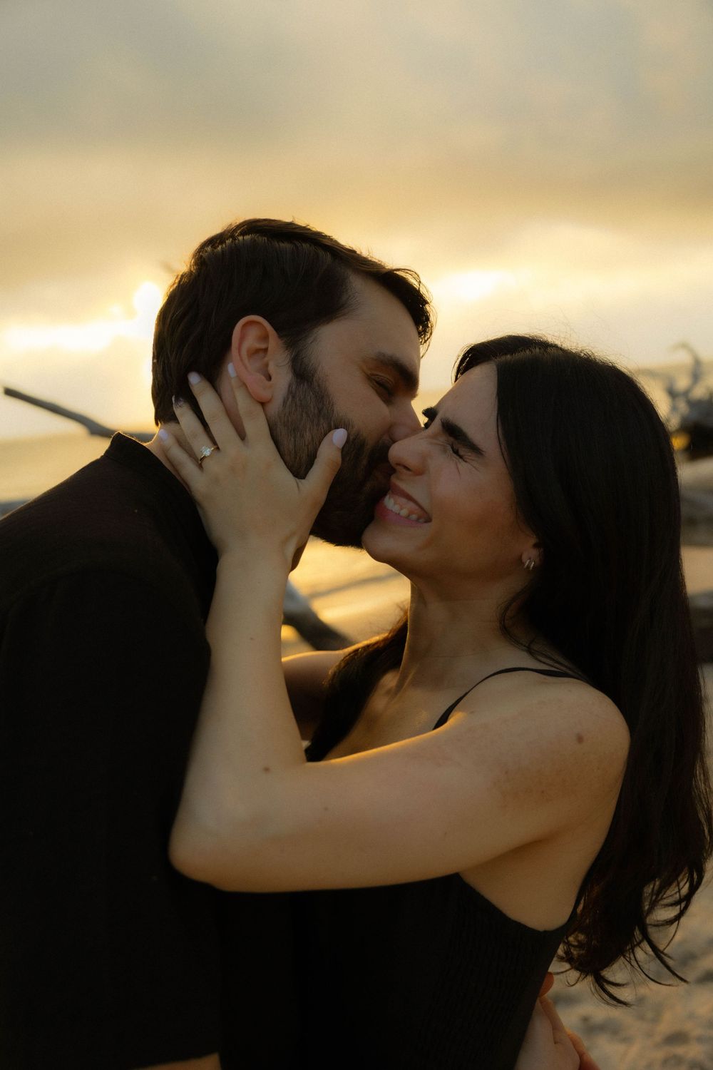 Couple sharing a romantic moment at sunset on the beach, woman smiling with engagement ring visible.