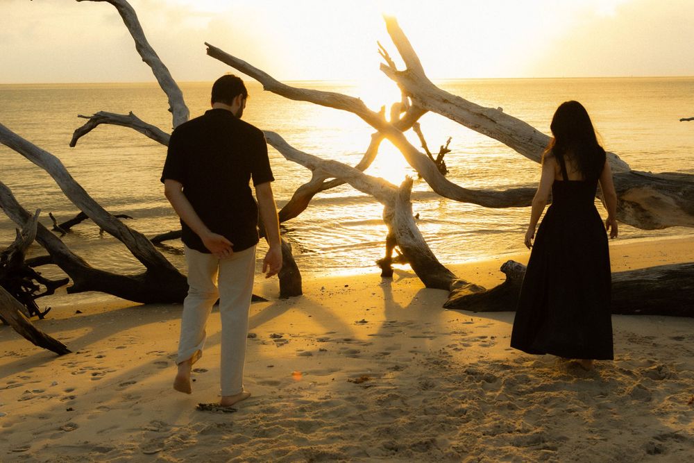 Couple standing near driftwood on a sandy beach at golden sunset by the ocean.