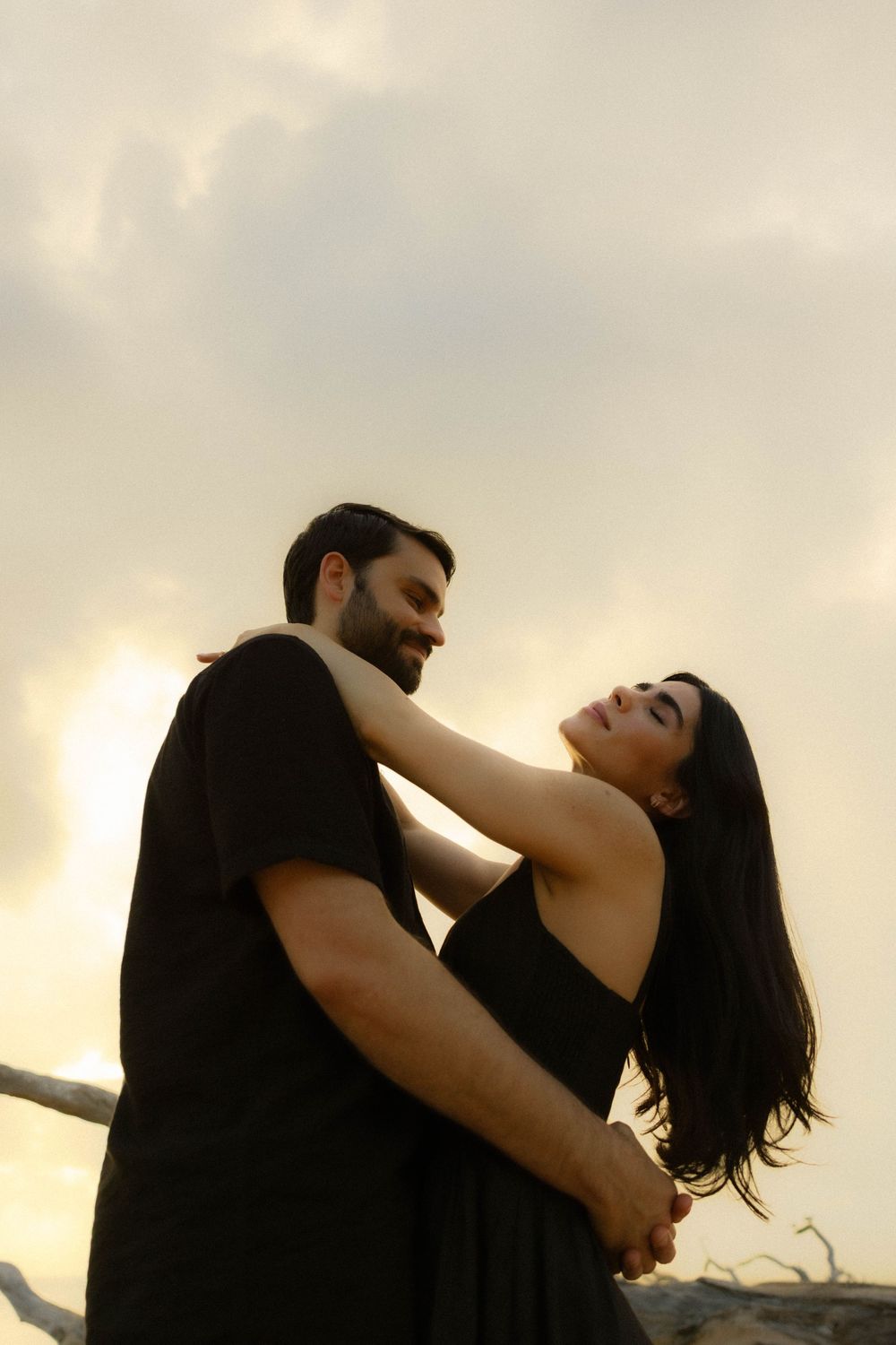 Romantic couple in black clothing dancing together against a dramatic sunset sky on the beach.