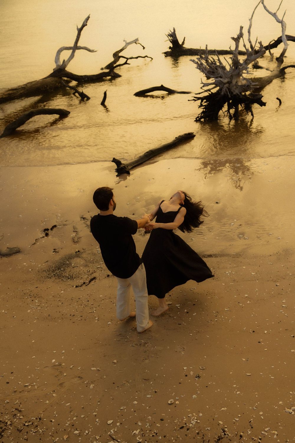 Couple dancing in black outfits on sandy beach surrounded by driftwood at sunset.