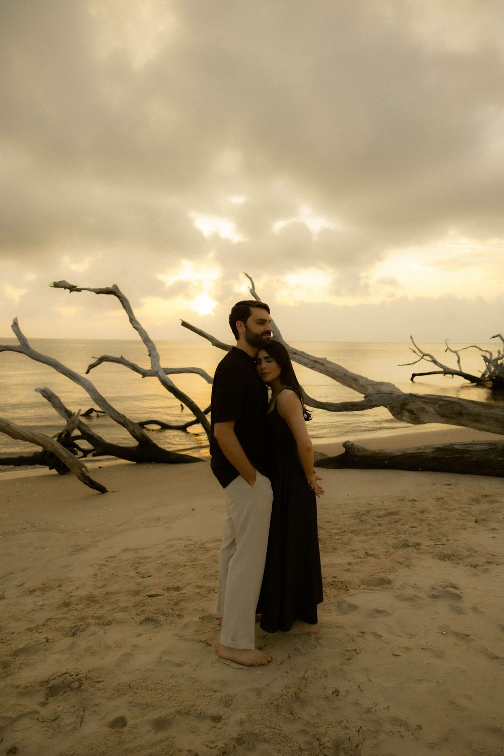 Couple embracing on sandy beach at sunset with driftwood and dramatic cloudy sky in background.