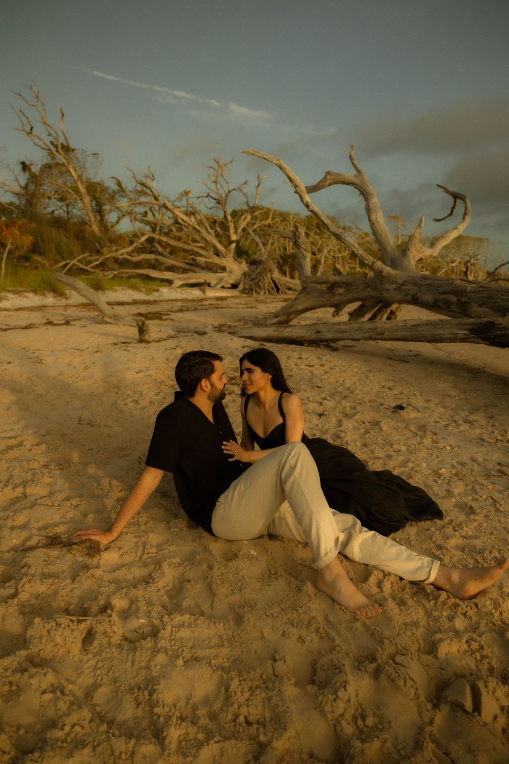 Couple sitting intimately on sandy beach at golden hour with driftwood trees in background.
