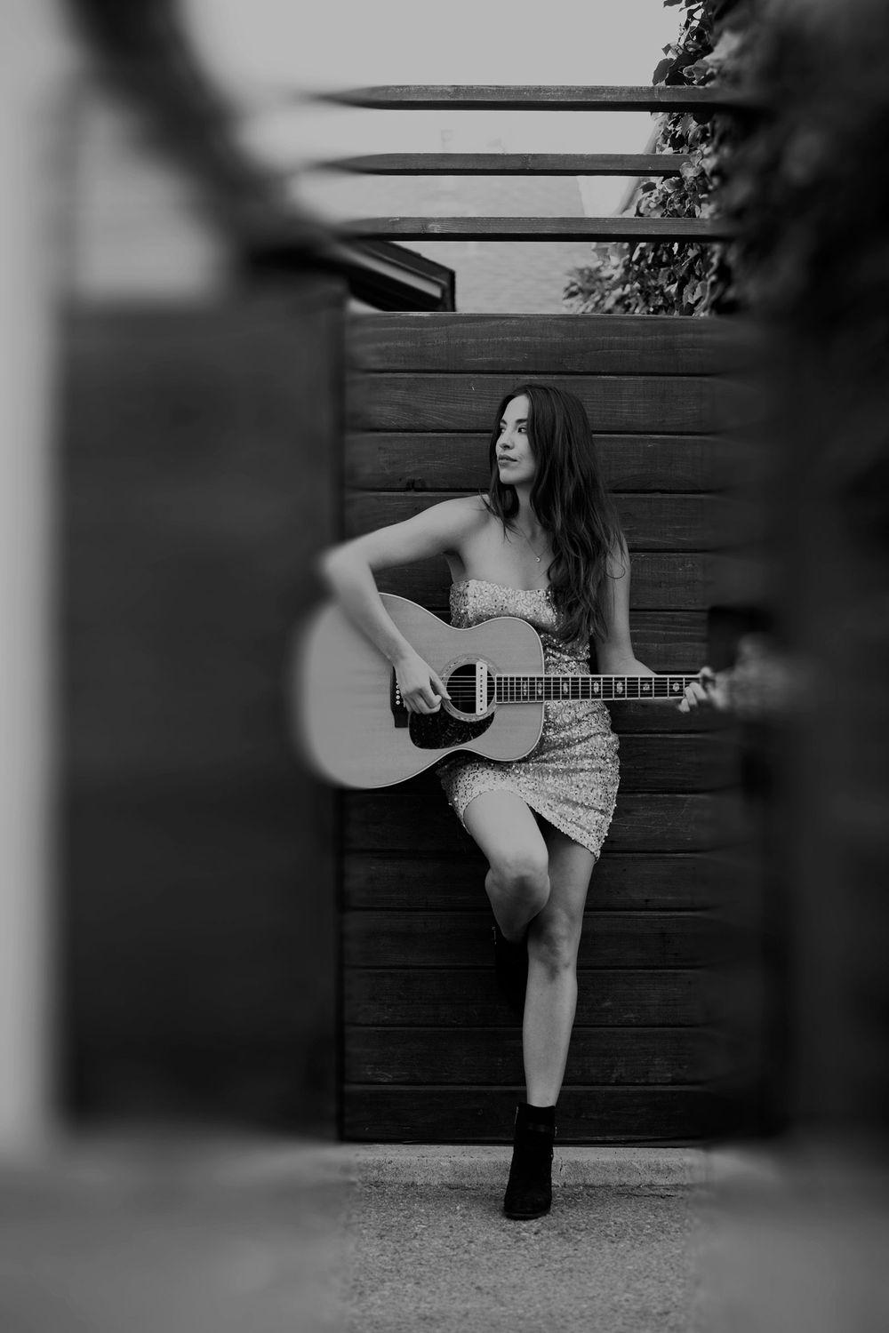 Black and white photo of woman in sequin dress playing acoustic guitar by wooden fence.