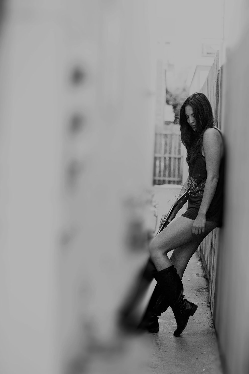 Black and white photo of woman in boots leaning against wall in narrow alleyway.