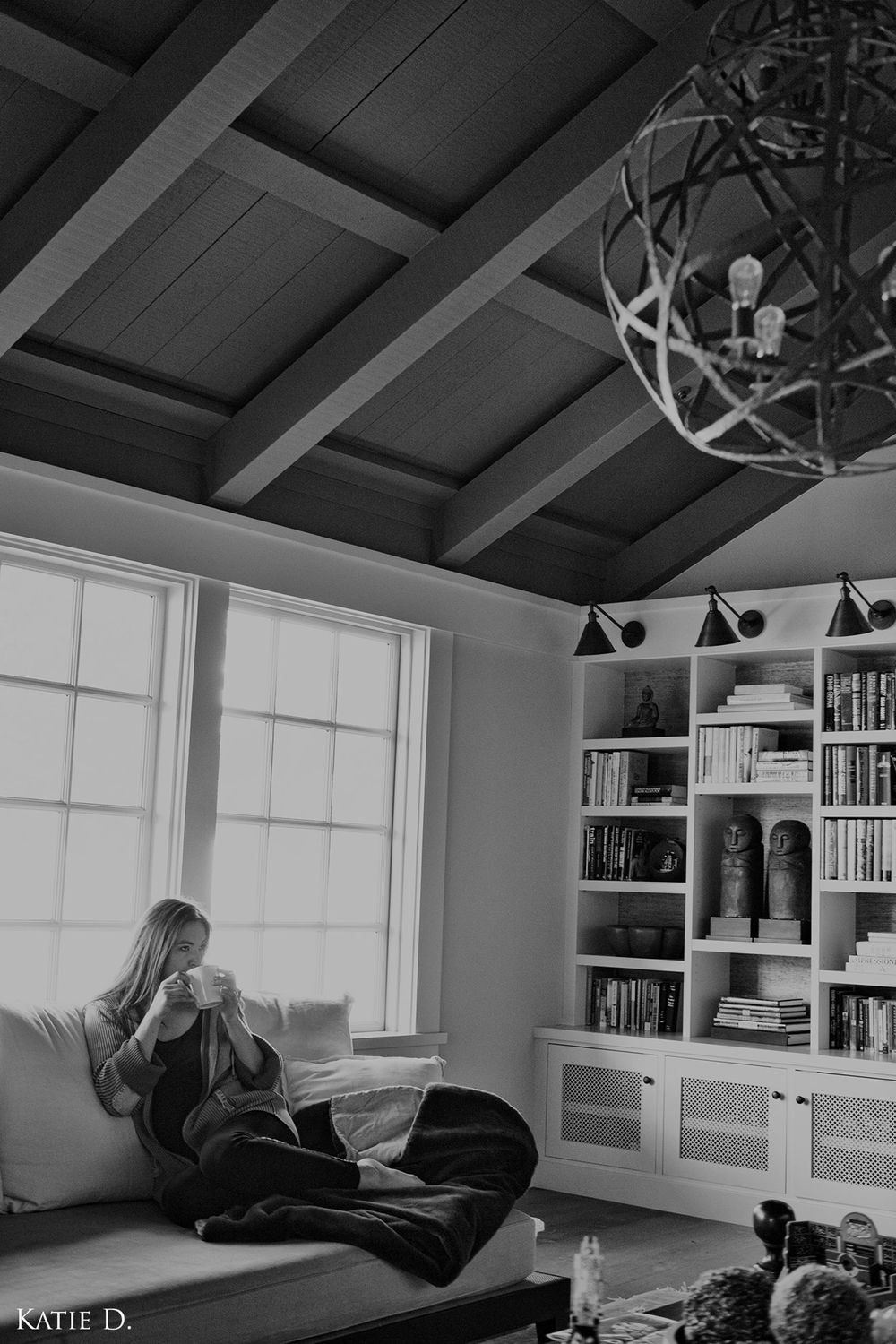 Black and white photo of a woman reading on a couch by a window in a room with built-in bookshelves.