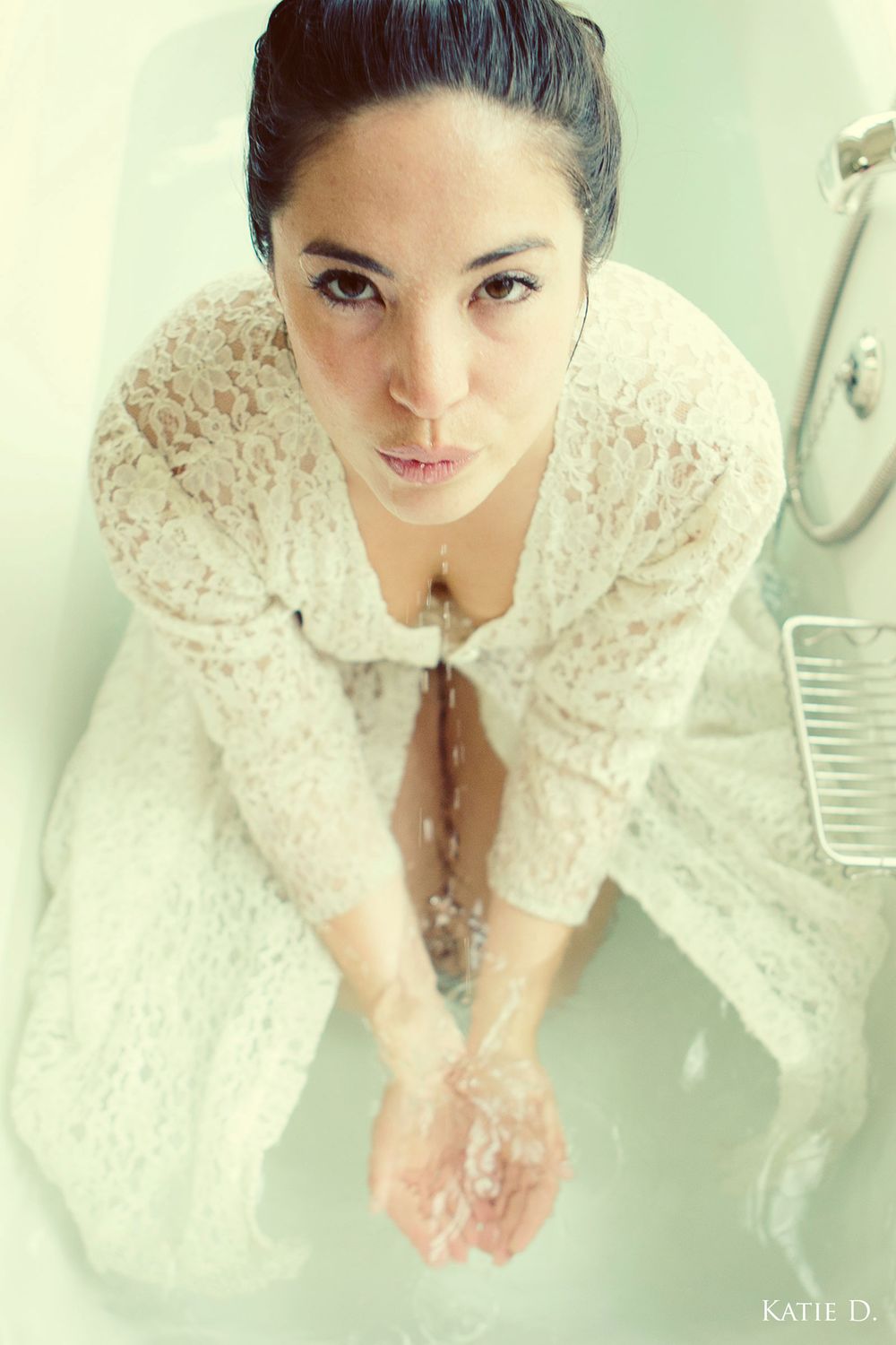 Overhead view of brunette woman in wet white lace robe sitting in bathtub cupping water in hands.
