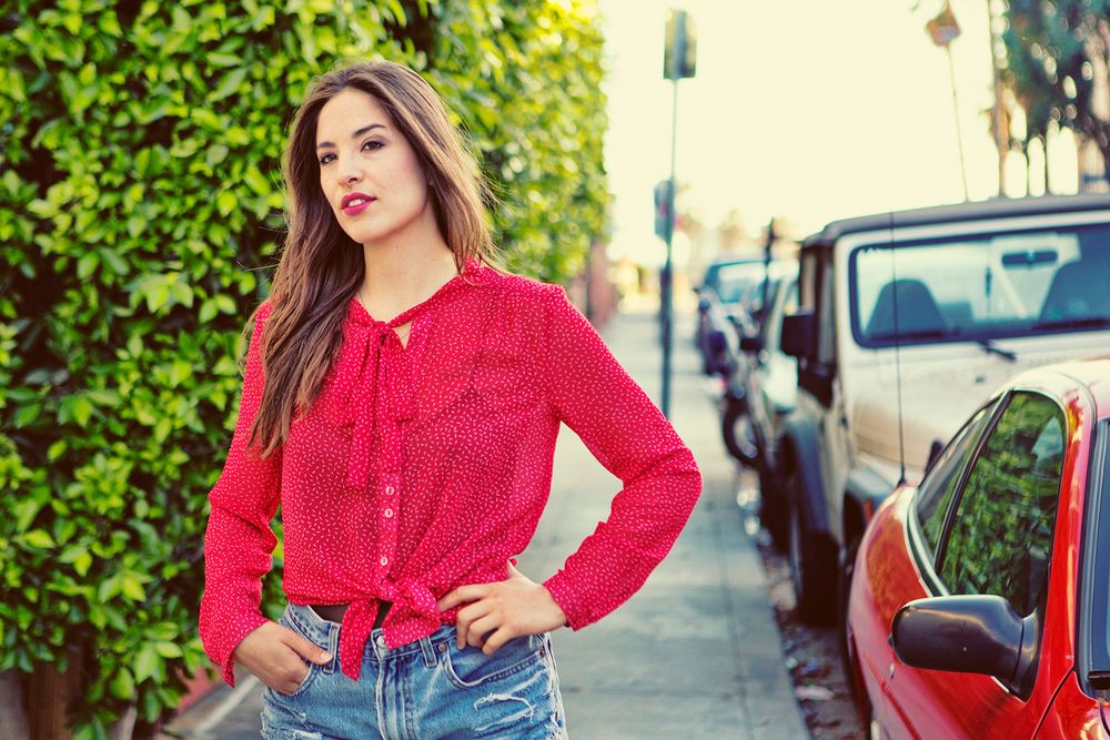 Young woman in red tied blouse and denim shorts posing on sunny urban sidewalk near parked cars.