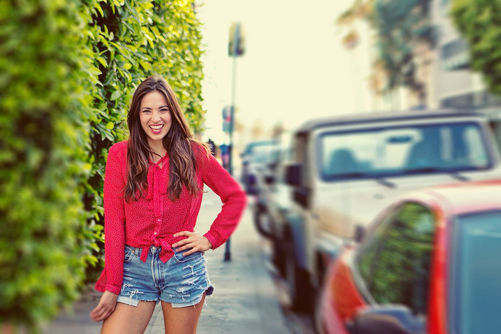 Cheerful young woman in red top and denim shorts laughing on a city sidewalk with parked cars in background.