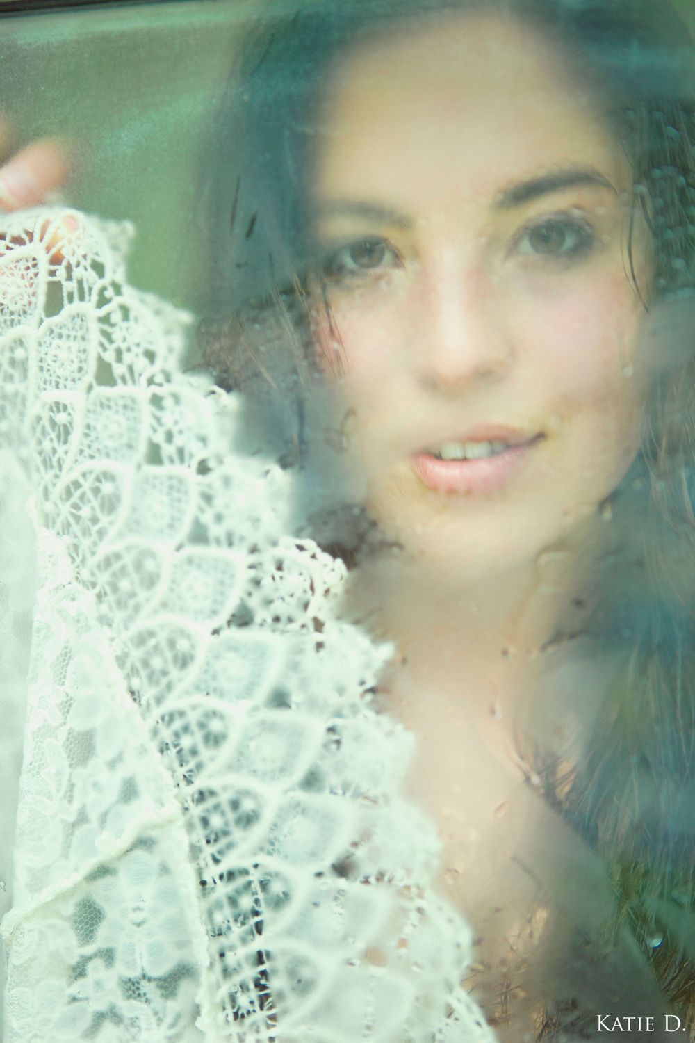 Dreamy close-up portrait of a young brunette woman holding white lace fabric, shot through a misty window.