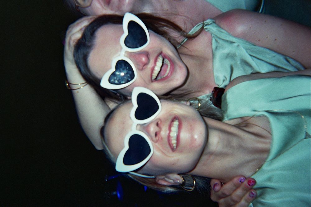 Two women wearing white heart-shaped sunglasses smile and pose together at a party.