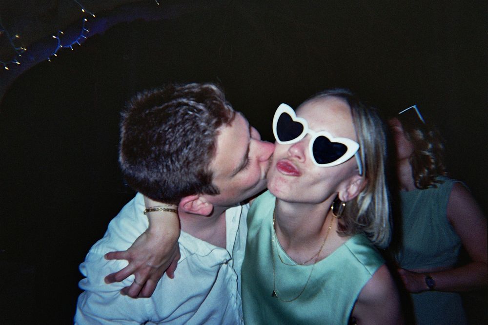 A man kisses a woman wearing white heart-shaped sunglasses at a party.