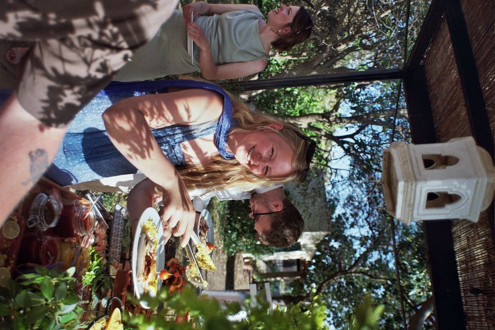 A young blonde woman smiles while gardening outdoors, surrounded by lush green plants and trees on a sunny day.
