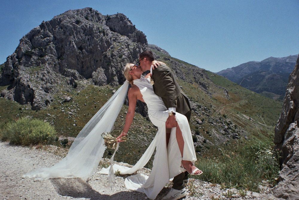 Bride and groom share a romantic dip kiss on a mountain trail, with dramatic rocky peaks and green valleys behind them.