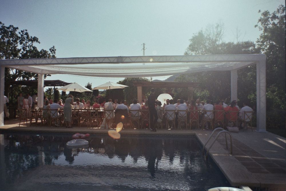 Guests gathered poolside under a white pergola canopy at an outdoor party on a sunny day.