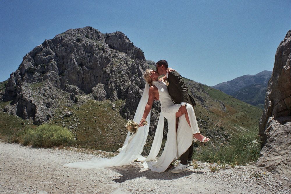A groom dips his bride in a white wedding dress against a stunning rocky mountain landscape on a sunny day.
