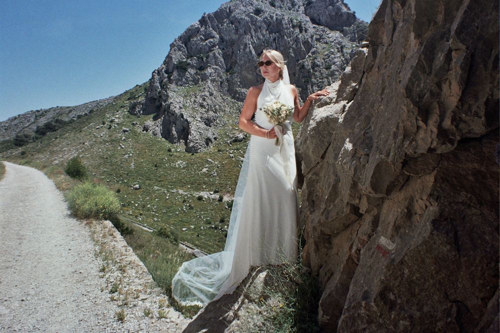 Bride in white gown poses against rocky mountain cliff on scenic winding road under clear blue sky.