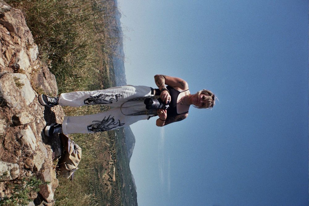 A hiker stands on a rocky mountain trail with a scenic valley and blue sky in the background.