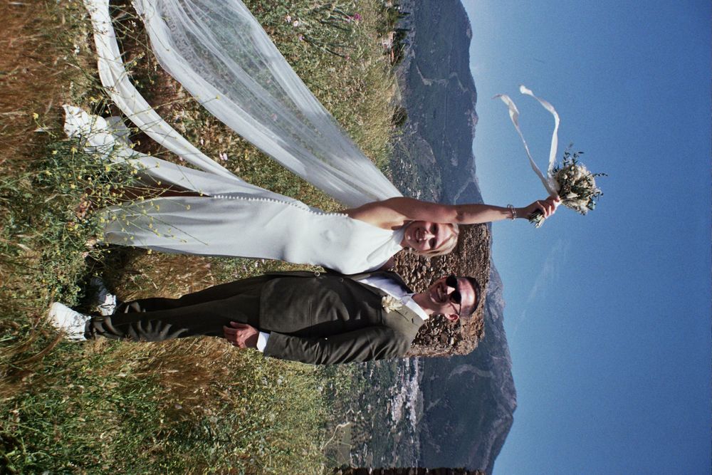 Bride and groom celebrating outdoors on a mountain hilltop, with veil flowing in the wind and bouquet raised.