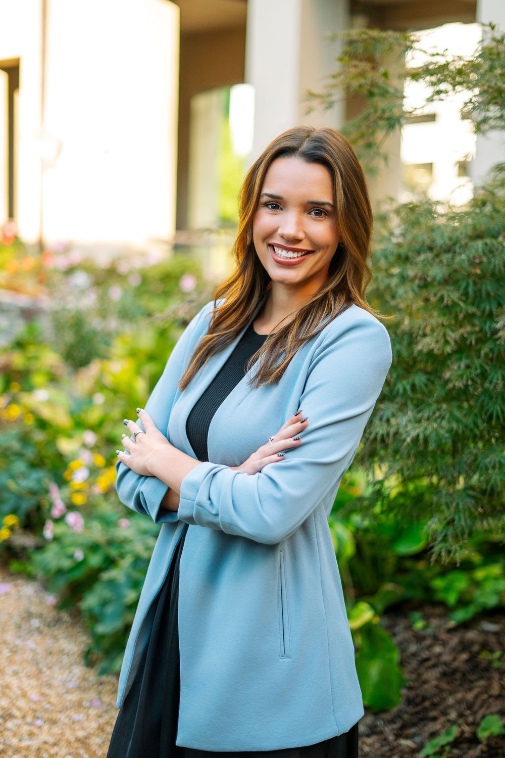 Professional young woman in light blue blazer smiling with arms crossed in garden setting.