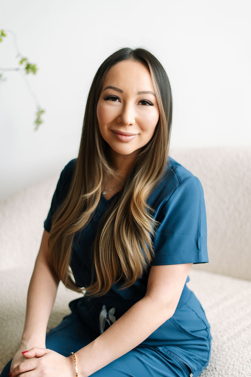 Female healthcare professional in blue scrubs sitting on a cream sofa, smiling warmly at camera.