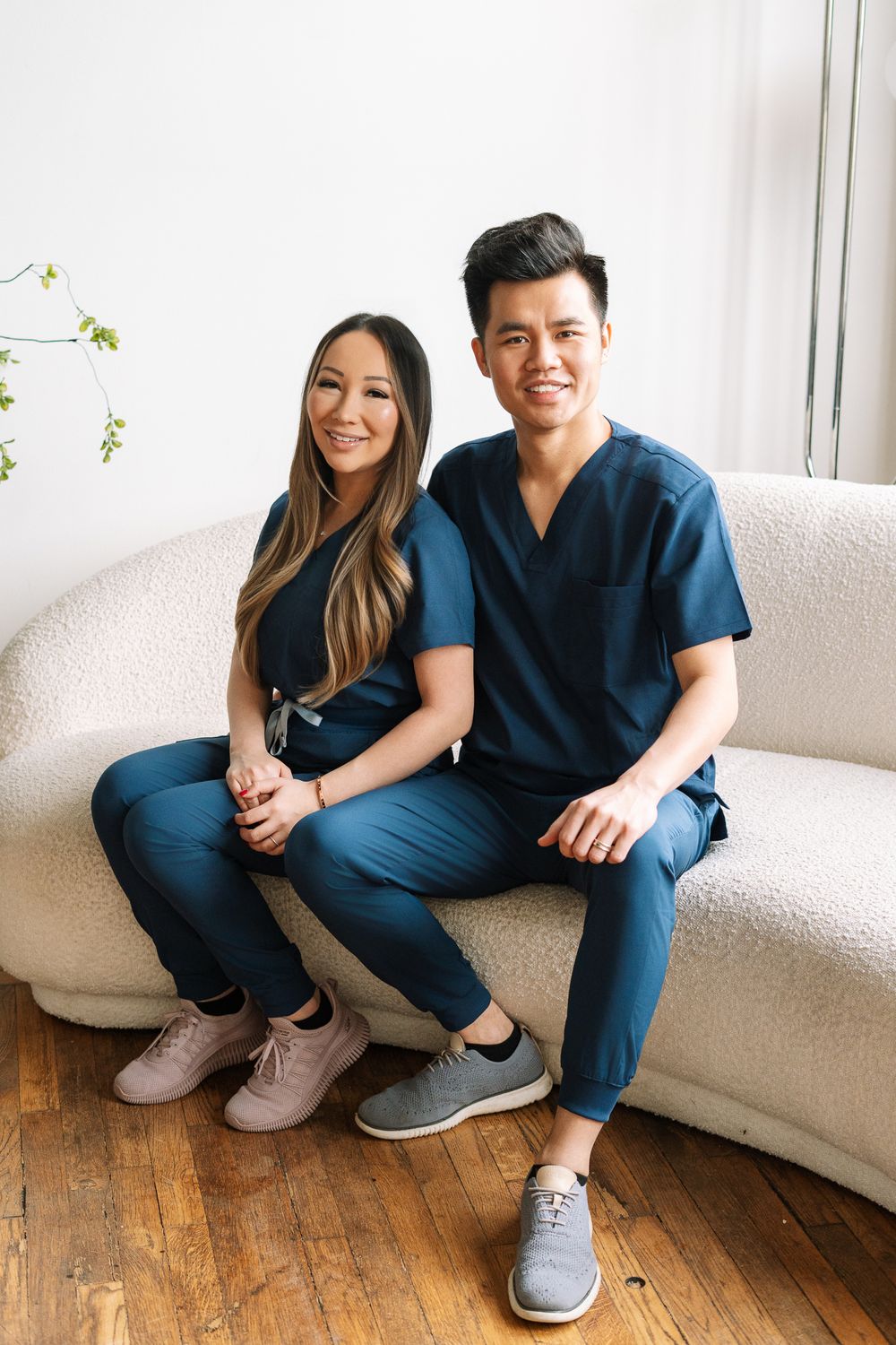 Two healthcare professionals in matching blue scrubs sitting together on a white sofa, smiling.