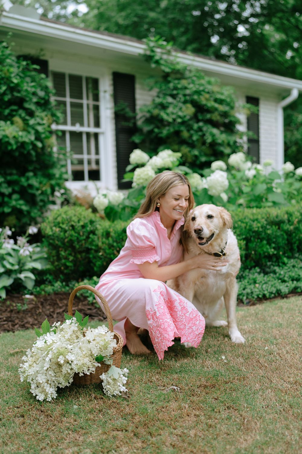 Smiling blonde woman in pink dress cuddling a golden retriever dog in a garden with hydrangeas.