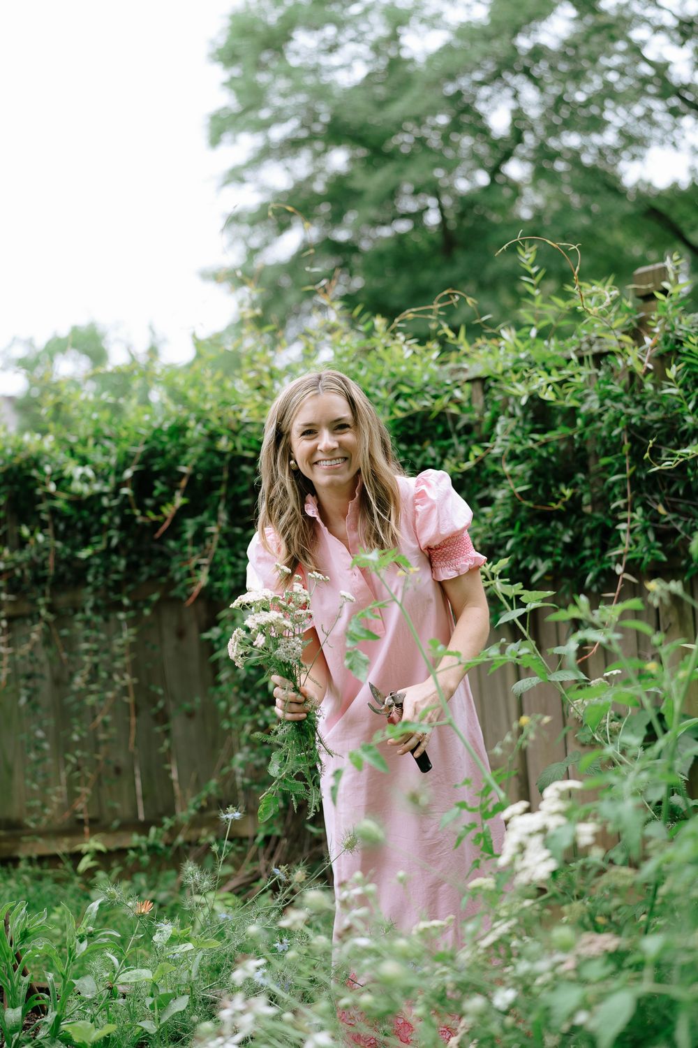 Smiling woman in pink dress holding wildflowers in a lush green garden.