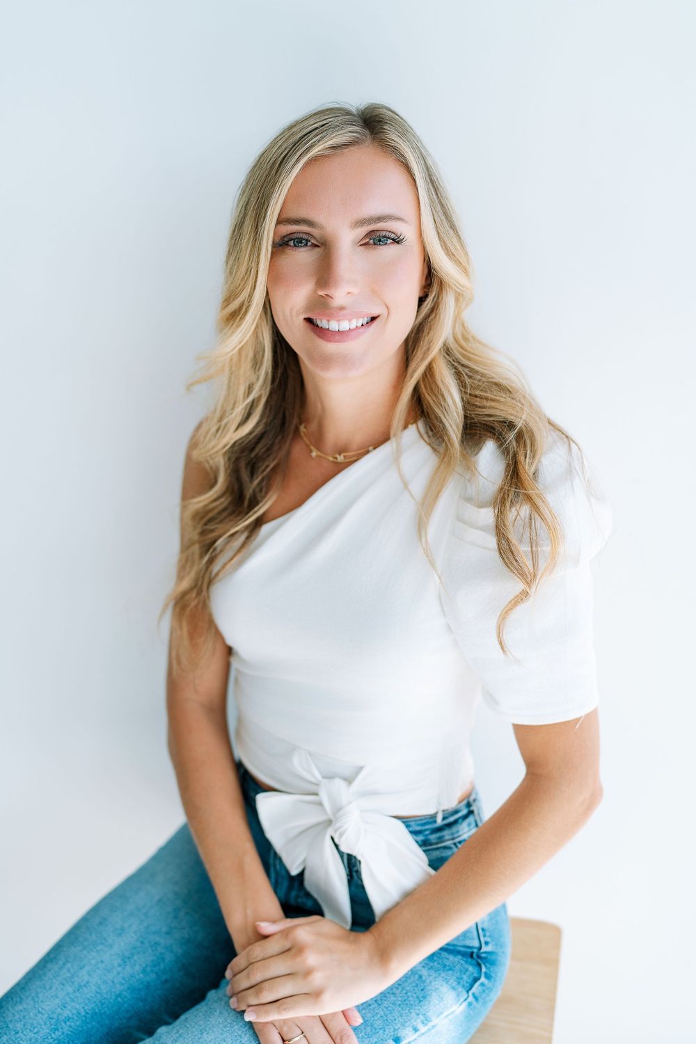 Smiling blonde woman in white one-shoulder top and jeans sitting against a clean white background.