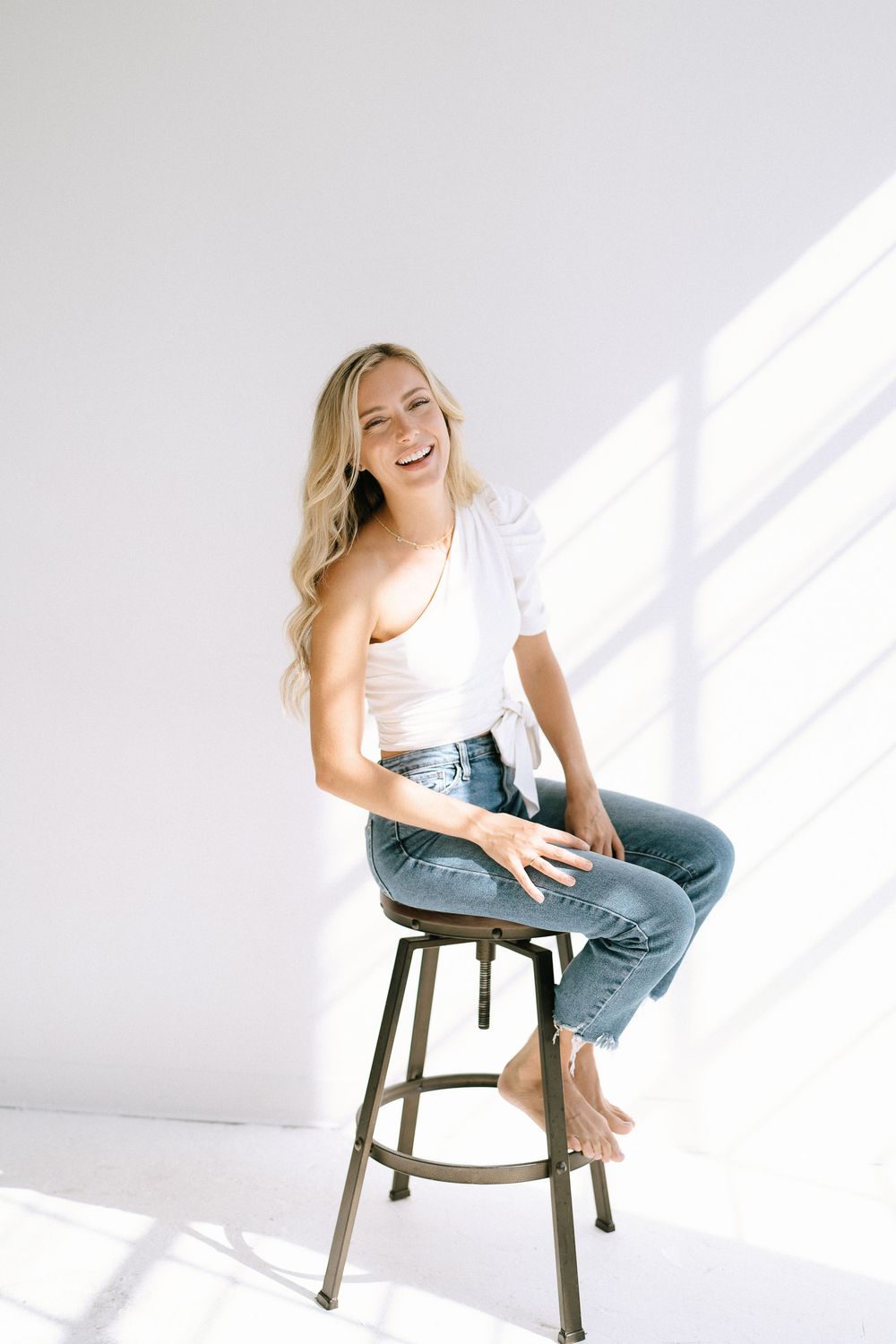 Laughing blonde woman in white top and jeans sitting barefoot on a stool in a bright studio.