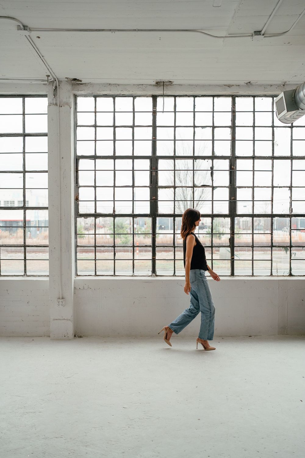A woman in a black top and light blue jeans walks barefoot across an empty industrial loft with large grid windows.