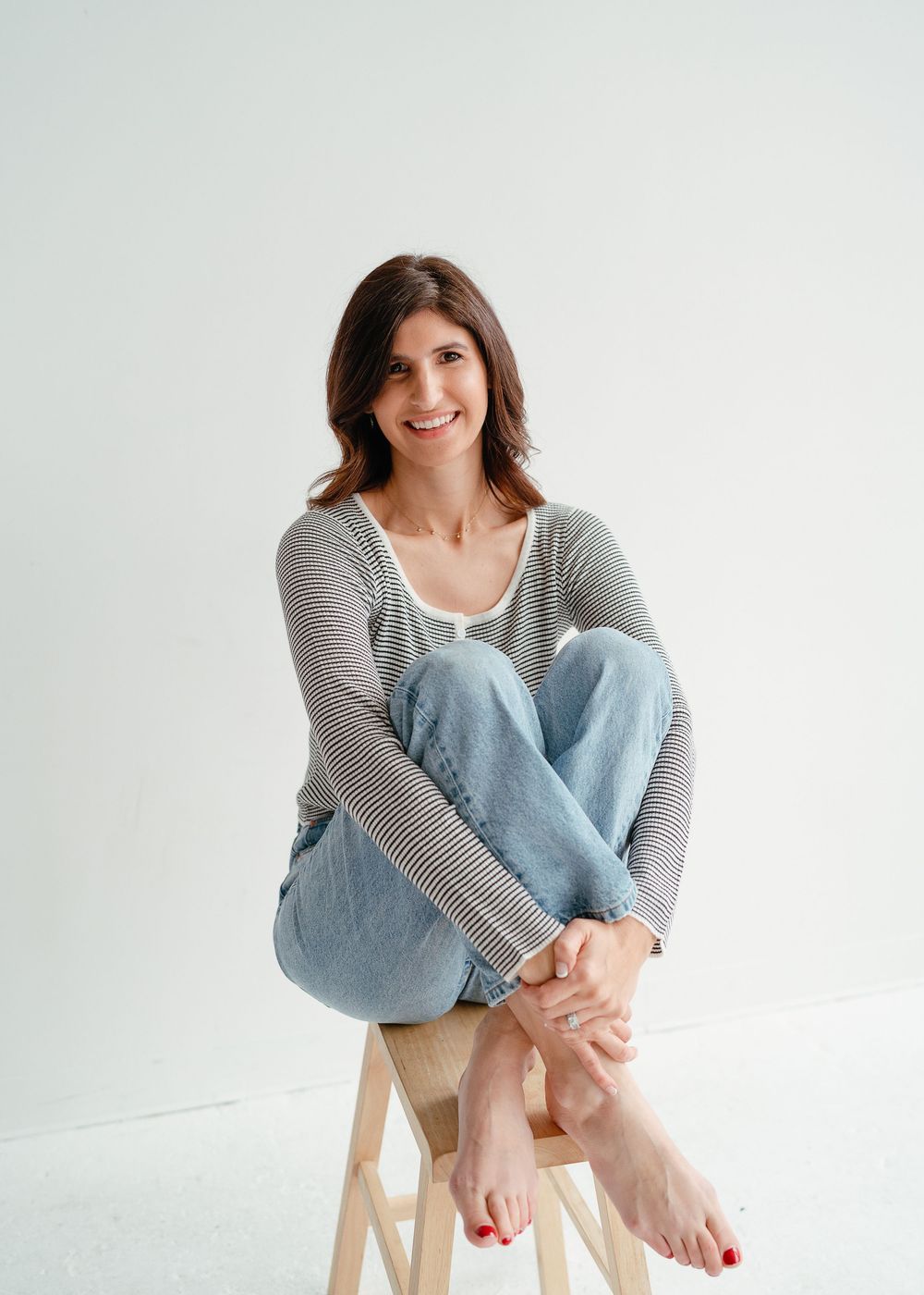 A smiling brunette woman sits cross-legged on a wooden stool wearing a striped long-sleeve shirt and light blue jeans.