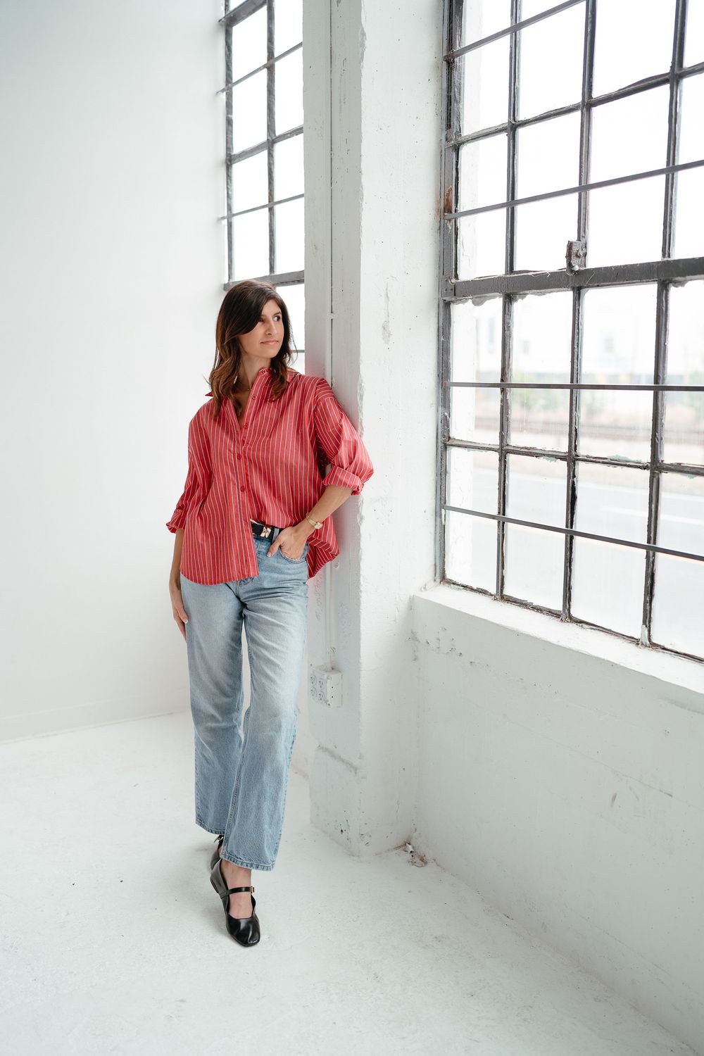 A woman in a red striped button-up shirt and light blue jeans leans against a wall near large industrial windows.